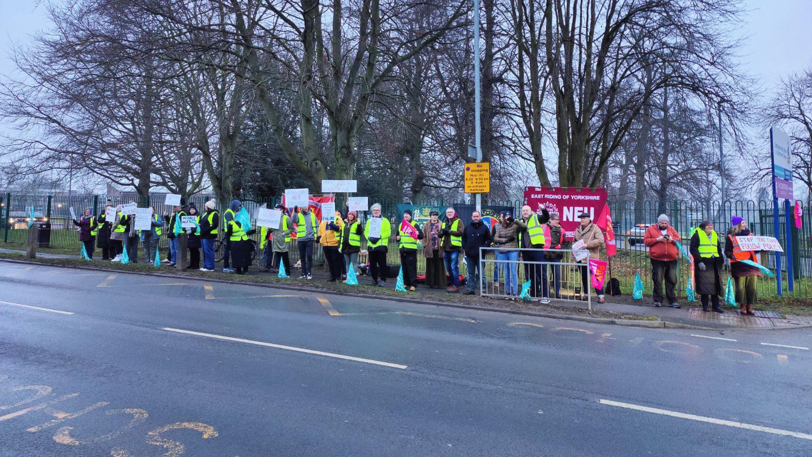 A row of people wearing hi-vis jackets are seen across an empty road on a picket line. They are stood in front of banners, and many are holding white signs.