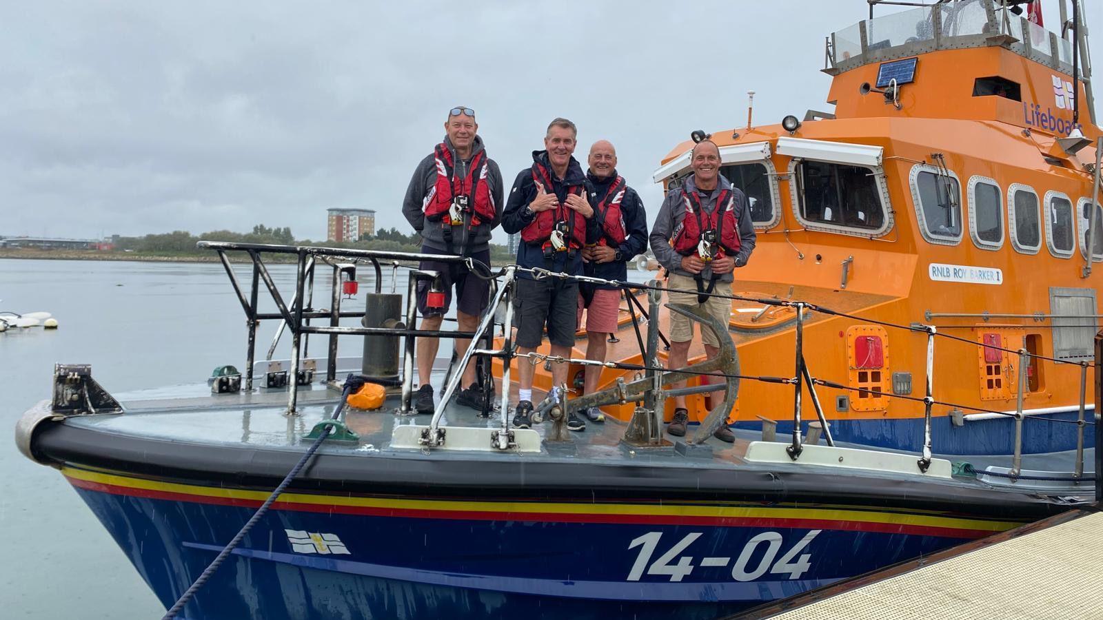 Four RNLI crew stand on the deck of an orange and blue all-weather Trent Class lifeboat.