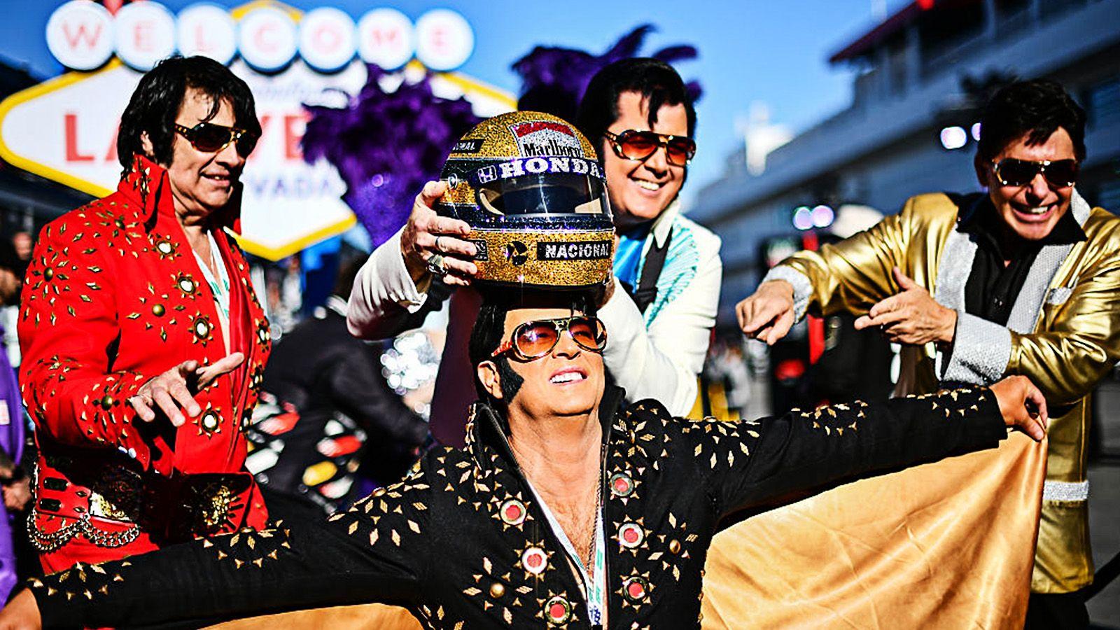 Four Elvis impersonators in vibrant, glittering outfits pose playfully near the Las Vegas sign, lifting a decorated racing helmet as they smile and strike dramatic stances in the bright sunlight.