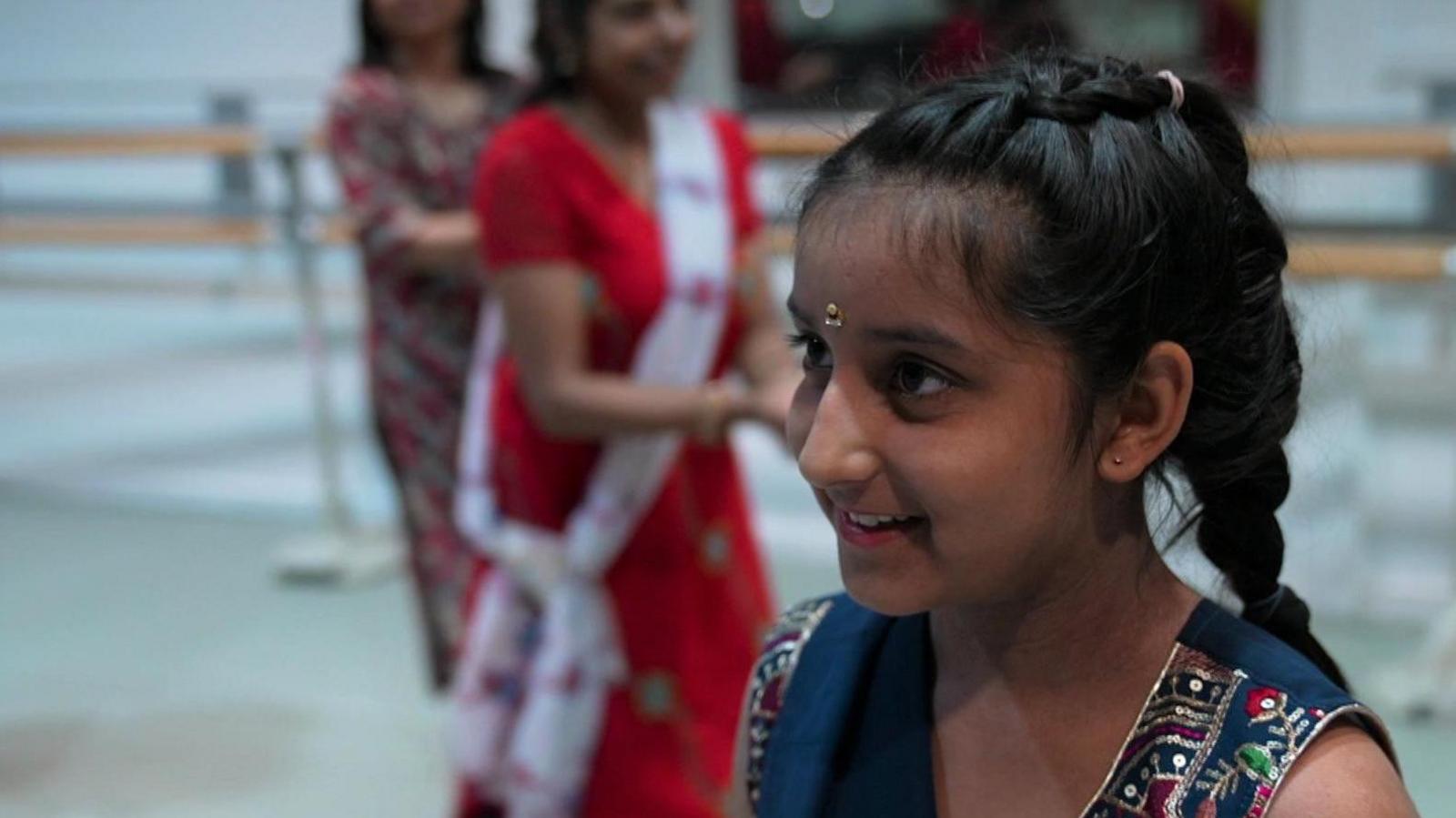 Young girl with long plaited hair wearing traditional Indian dress with blurred figures dancing in the background