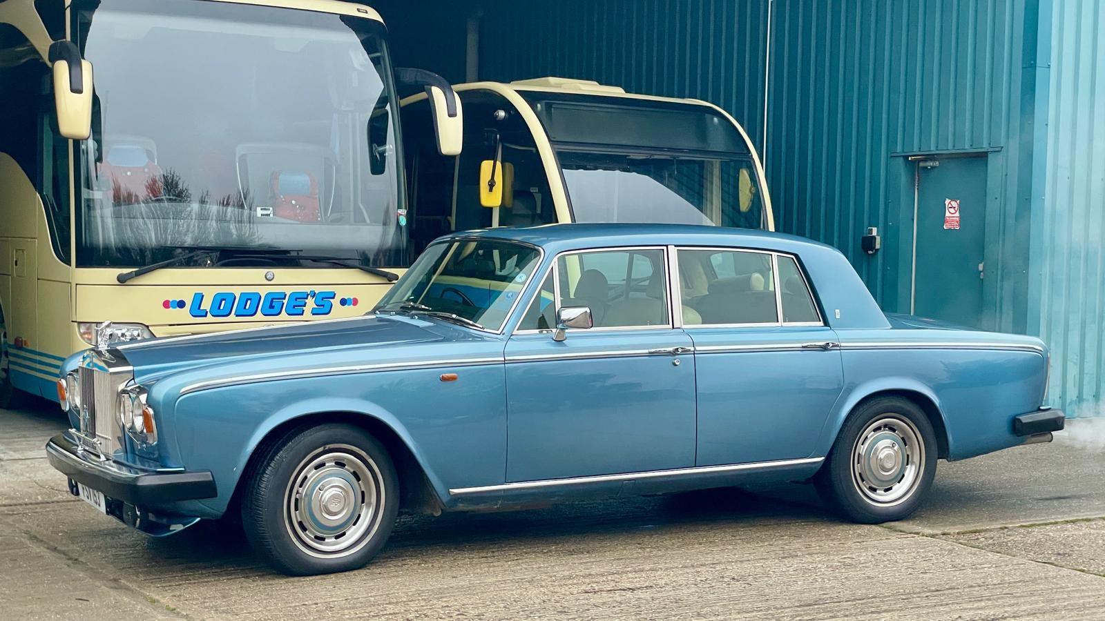 The blue Rolls Royce is parked in a car park in front of two larger buses.