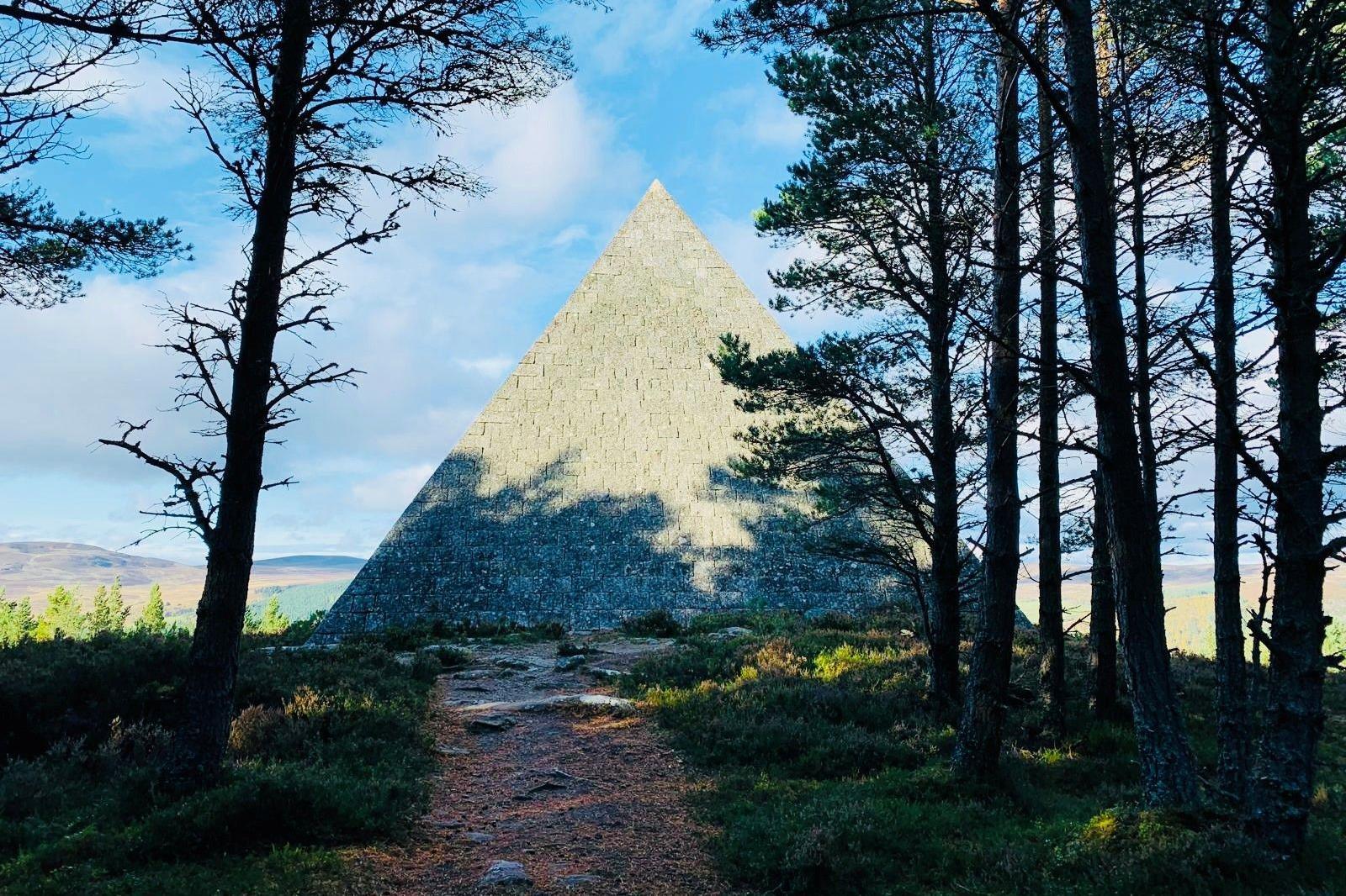 Pyramid-shape cairn at the edge of woodland, with the shadow of trees across the structure.