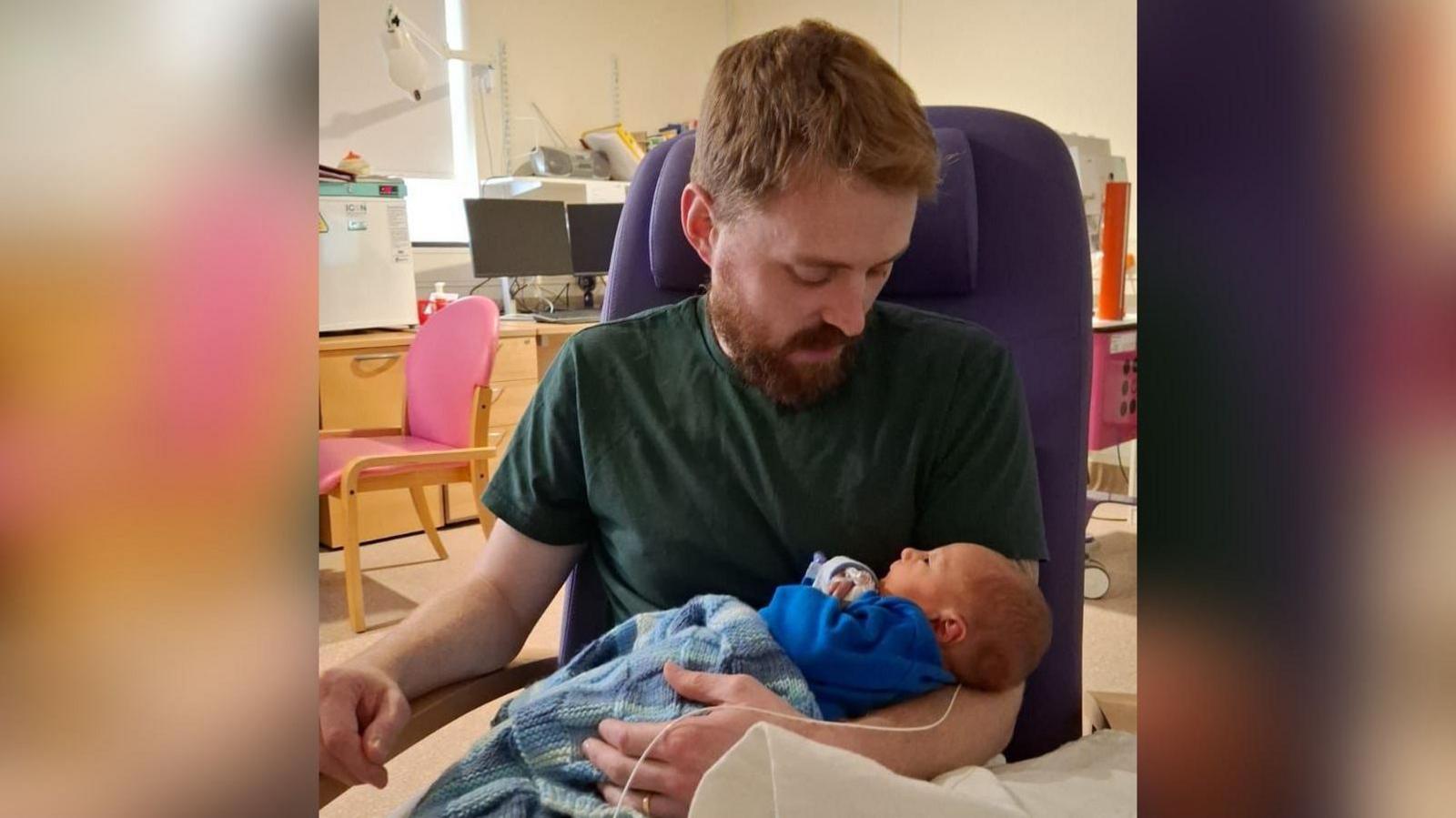 Lewis Daines in a hospital with his baby who is wrapped in a blue woollen blanket