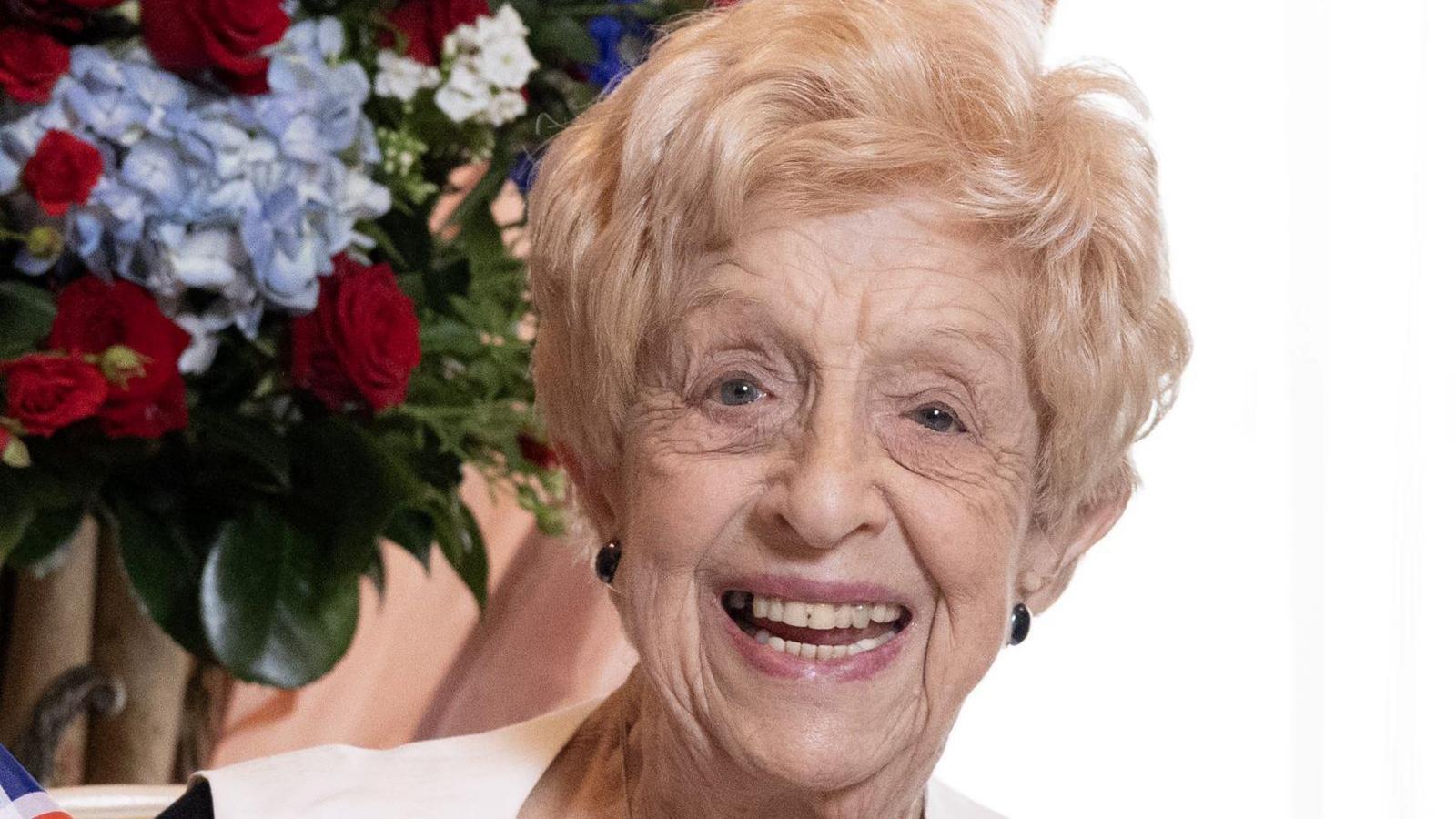 Ruth June Bourne has short blonde hair and is smiling in front of a bouquet of roses.