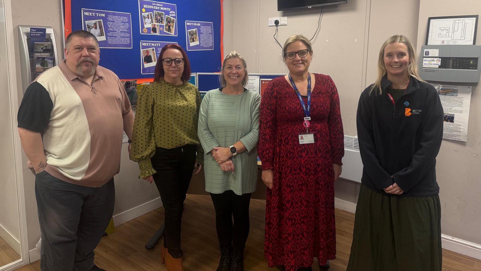 A man and four women stand in a line and smile as they look at the camera. They are in a room with laminate flooring and pale brown walls. Information boards are attached to the walls. On the far left, a man has cropped grey hair and a goatee beard and wears a brown and cream polo shirt. Next to him stands a woman with brown hair who wears glasses, a green polka-dot top and black jeans. In the centre of the line stands a woman with blonde hair, tied back, who wears a long light-green top over black leggings. Next to her stands a taller woman with blonde hair, tied back, who wears glasses, a long red and black patterned dress and a blue lanyard with white identity pass. On the far right stands a younger woman with blonde hair who wears a black fleece top and long black skirt.