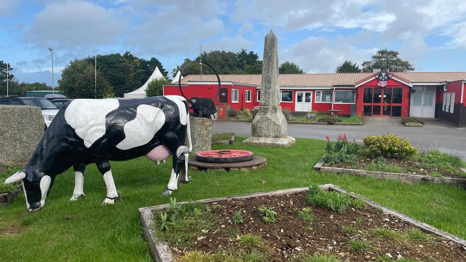 A model black and white cow on grass in front of red building, with a large wooden cow's head above the entrance