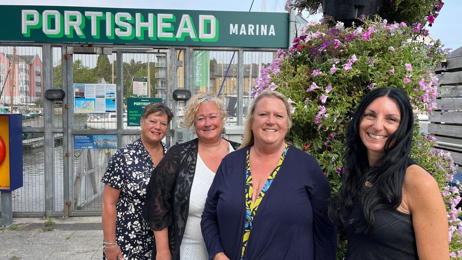 Four women stand in a line, slightly angled, facing the camera. They are all smiling and behind them is some fencing and an area. Also behind them is a large sign that says "Portishead Marina" in capital white letters on a green background.