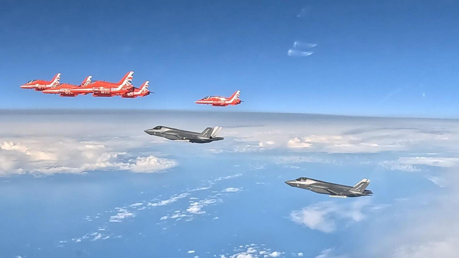 Five Red Arrows planes fly ahead of two F-35B aircraft, which are painted grey. You can see clouds and the sea below the aircraft. 