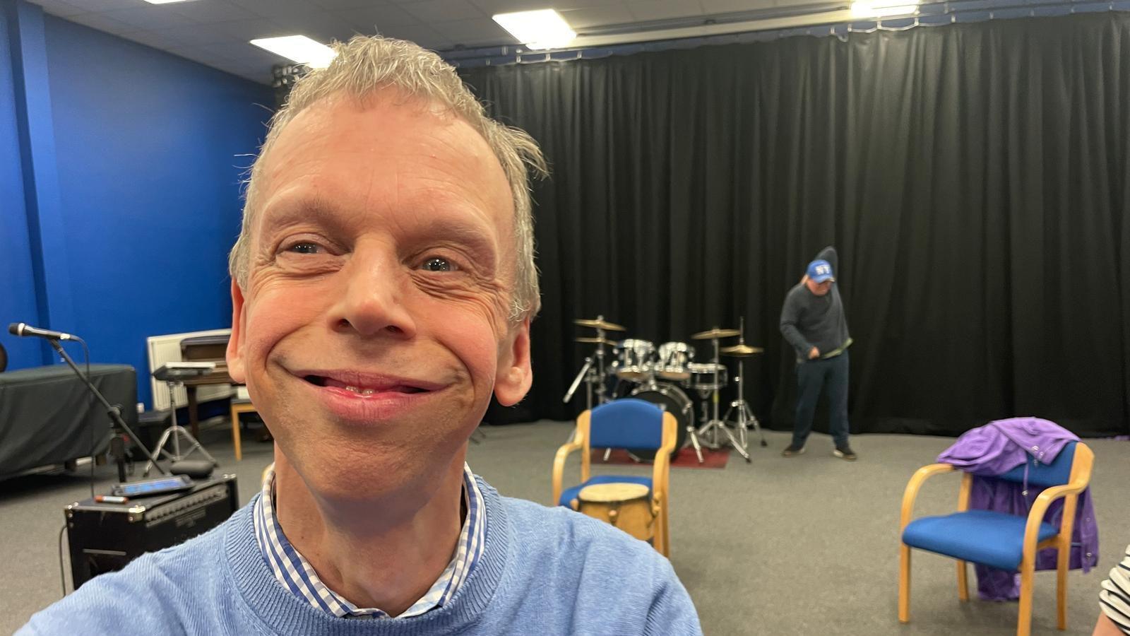 Paul is smiling wearing a chequered colour, which is blue and white and a light blue jumper on top. In the background there is two chairs, and various instruments dotted around.