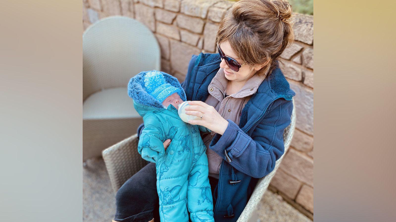 A woman sitting in a chair outside feeding a baby from a bottle. She had dark hair tied back and is wearing sunglasses, a blue coat with a light purple fleece underneath, and dark blue trousers. The baby is wearing a bright blue padded romper suit and bright blue woolly hat. There is a brick wall behind them.