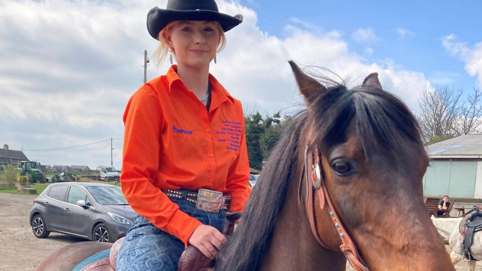 A teenage girl wearing a black cowboy hat and bright orange shirt sits astride a brown horse wearing a Western-style saddle.