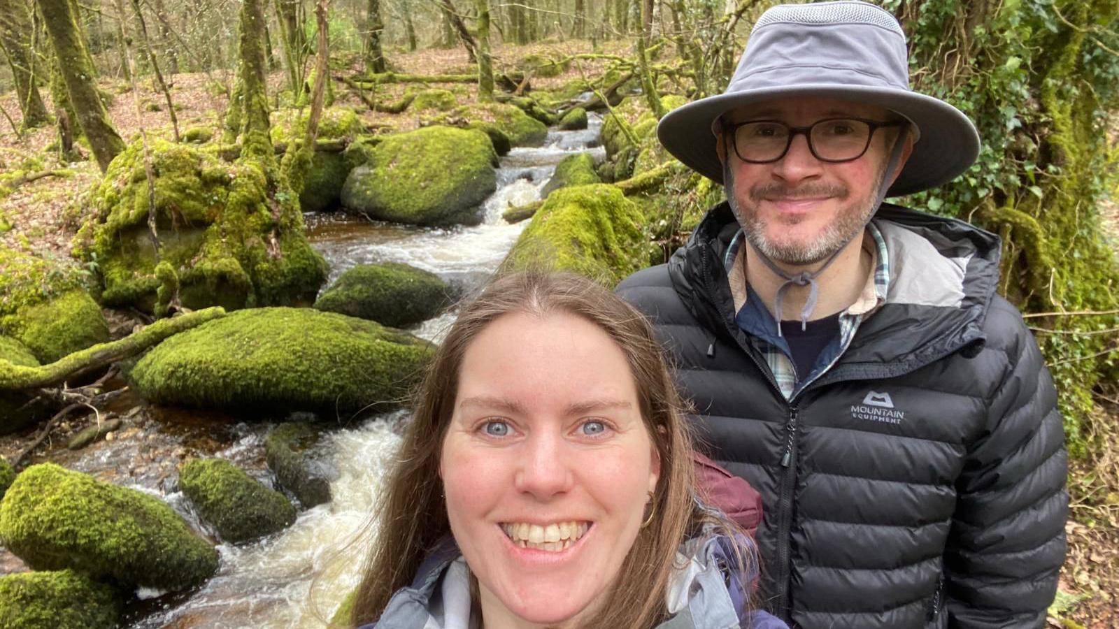 Holly Jackman and Gavin Robbins smile while taking a selfie on a walk near Manaton on Dartmoor. A stream is flowing behind them. Rocks in the area has moss on them. Gavin is wearing a large brimmed hat.