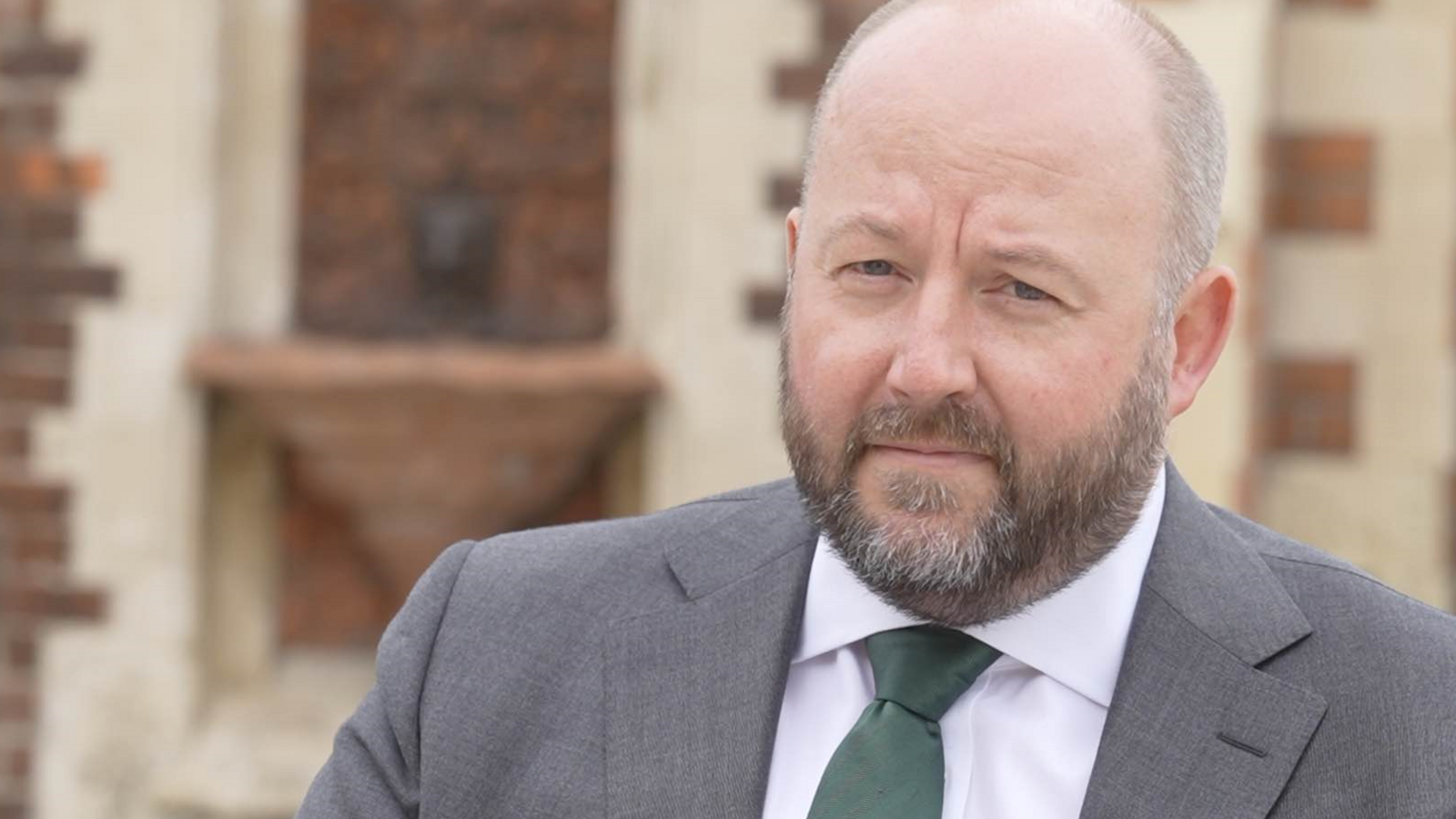 Nick Timothy looks at the camera as he photographed outside. He has facial hair and is wearing a grey suit with a green tie. Behind him is a red-bricked building. 