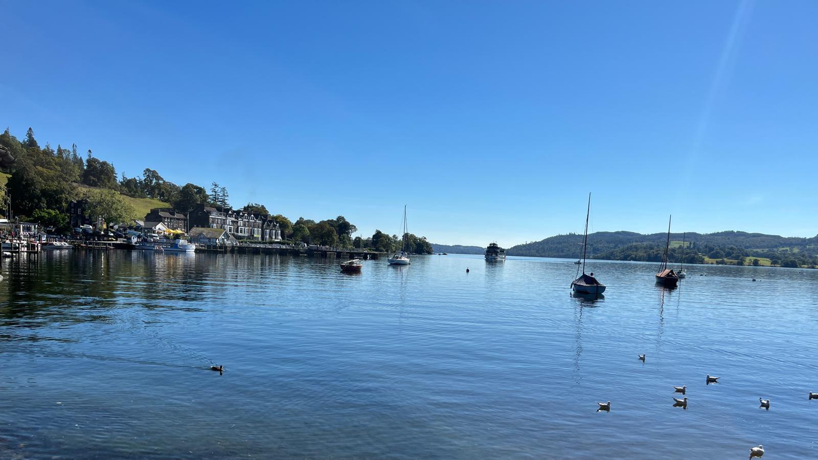 A view of Windermere on a sunny day with houses on one bank, hills in the distance and boats on the water.