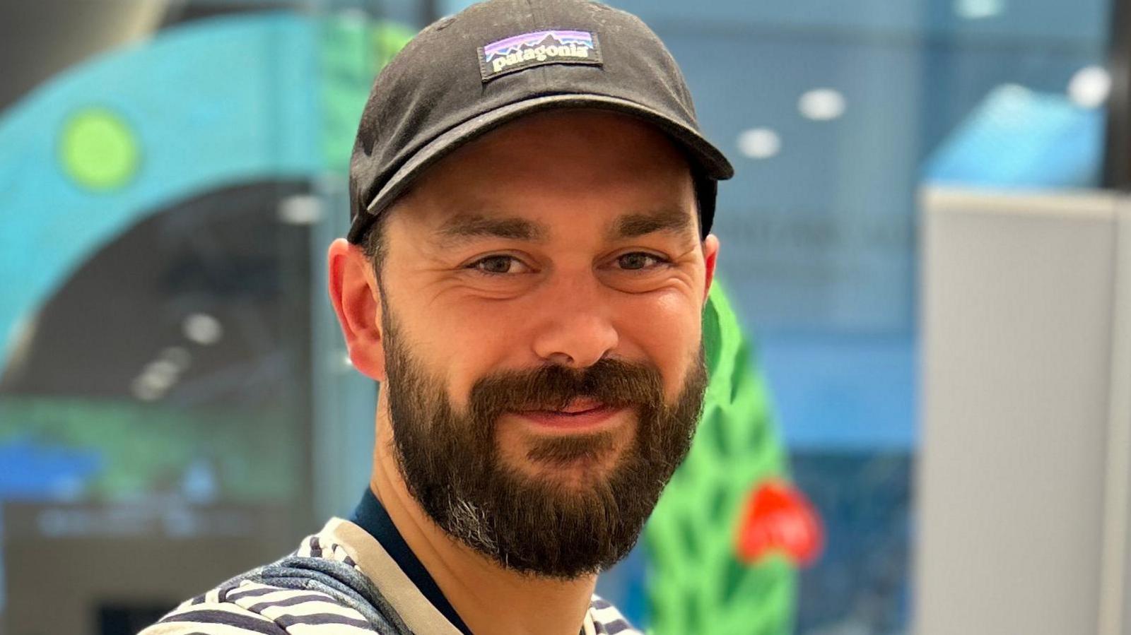 Adam is smiling at the camera while inside. His face is cast in a warm orange glow from the lighting. He has a blue and white stripy top on and a grey Patagonia cap. He has brown eyes and a dark brown beard. 