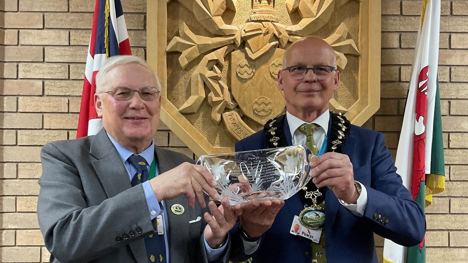 Gareth Morgan (left) stands next to Jonathan Wilkinson (right) holding a glass inscribed plague. Mr Morgan has silver hair and smiles at the camera. He wears a dark grey suit, pale blue shirt and navy tie. Mr Wilkinson also holds the plague and smiles at the camera. He wears a navy suit, white shirt and green tie.