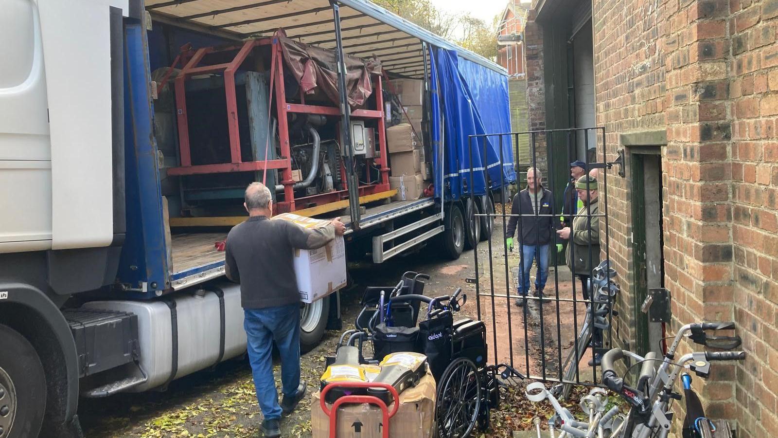 Men carry boxes to load  onto an articulated lorry. The curtain on the side of the lorry is open, showing a large electricity generator on the flatbed.