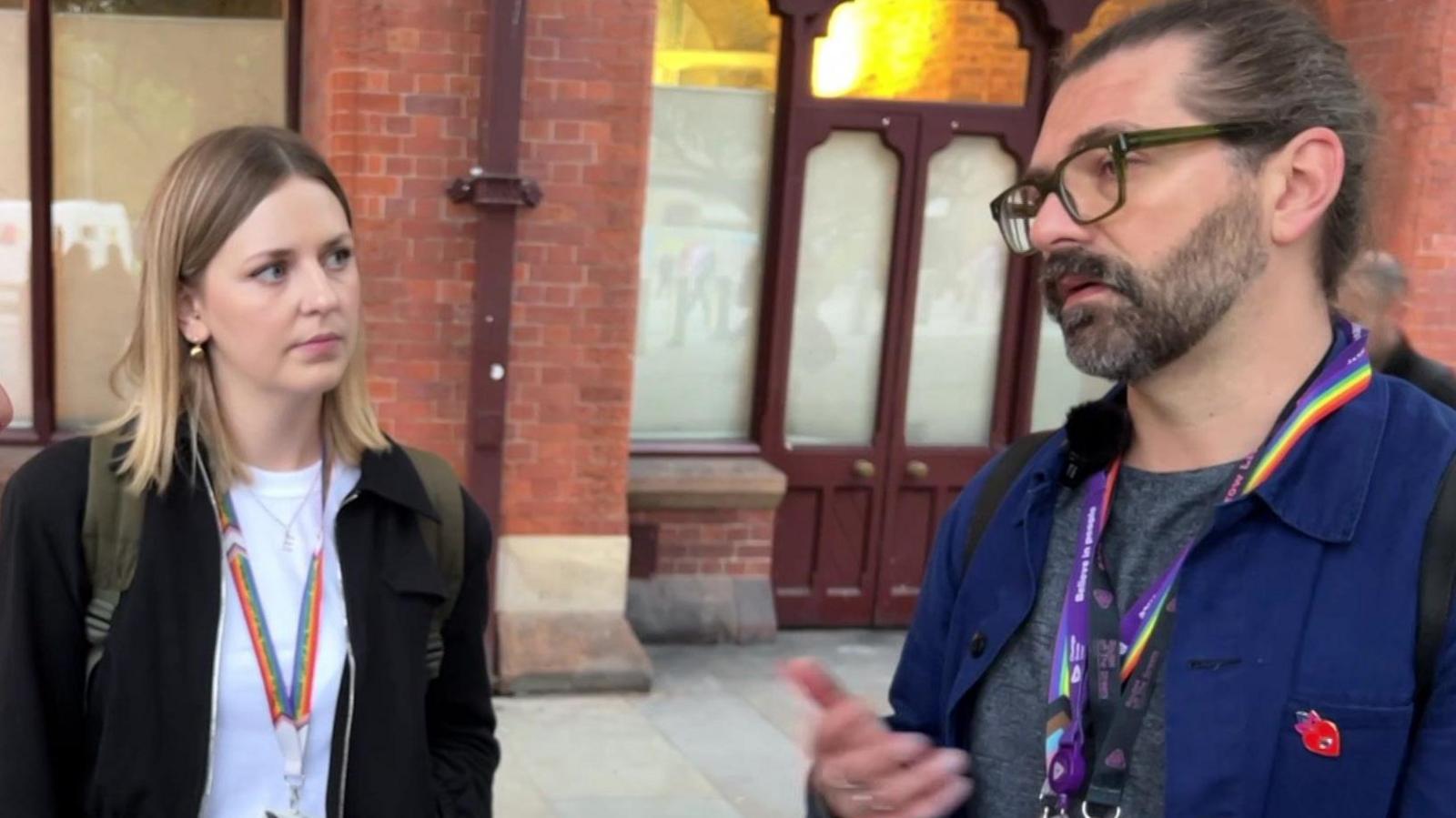 A man explains something to a third person, while standing next to a woman with blond, shoulder-length hair.

