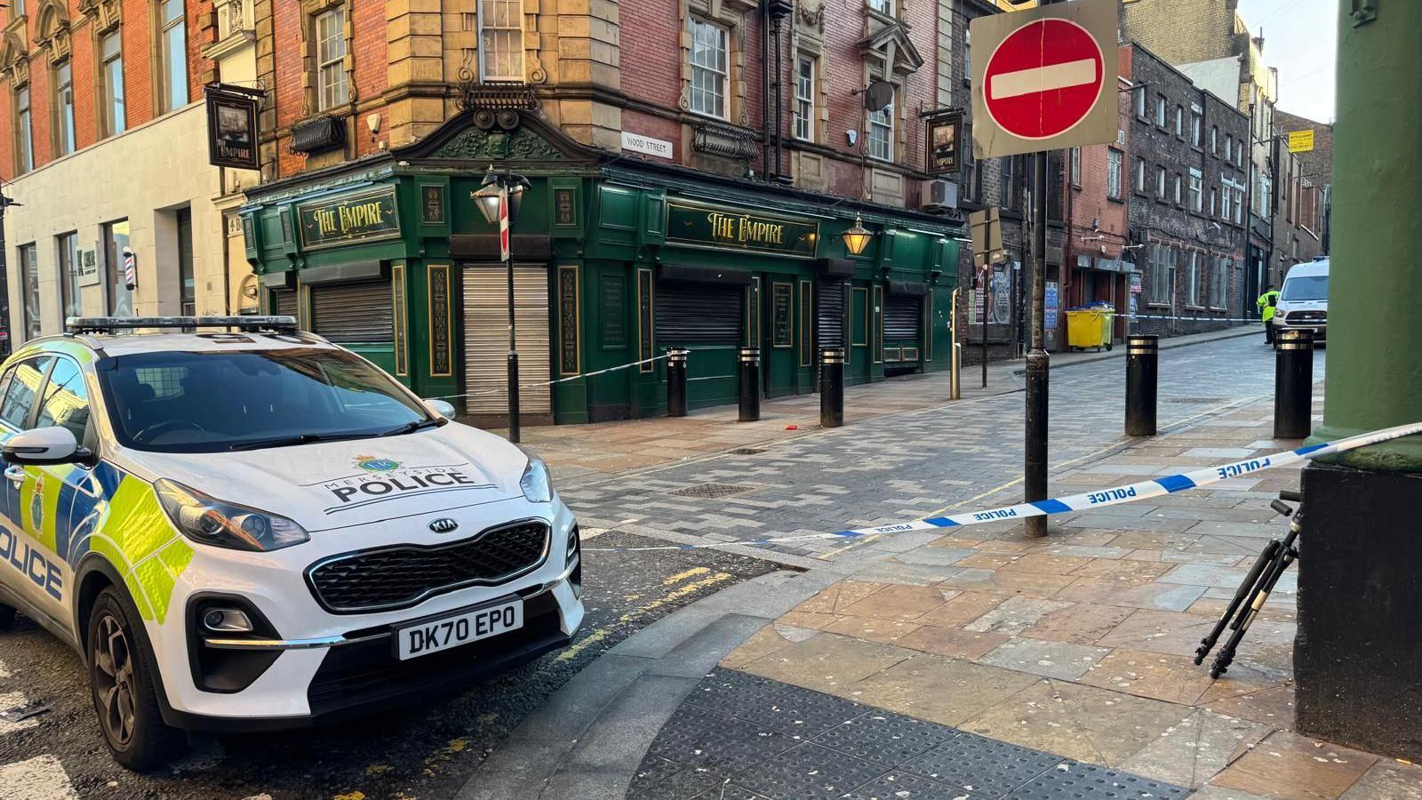 A police patrol car is parked at the mouth of a junction which is cordoned off by blue and white police tape. The front of a pub, with a sign saying The Empire, is in the backdrop