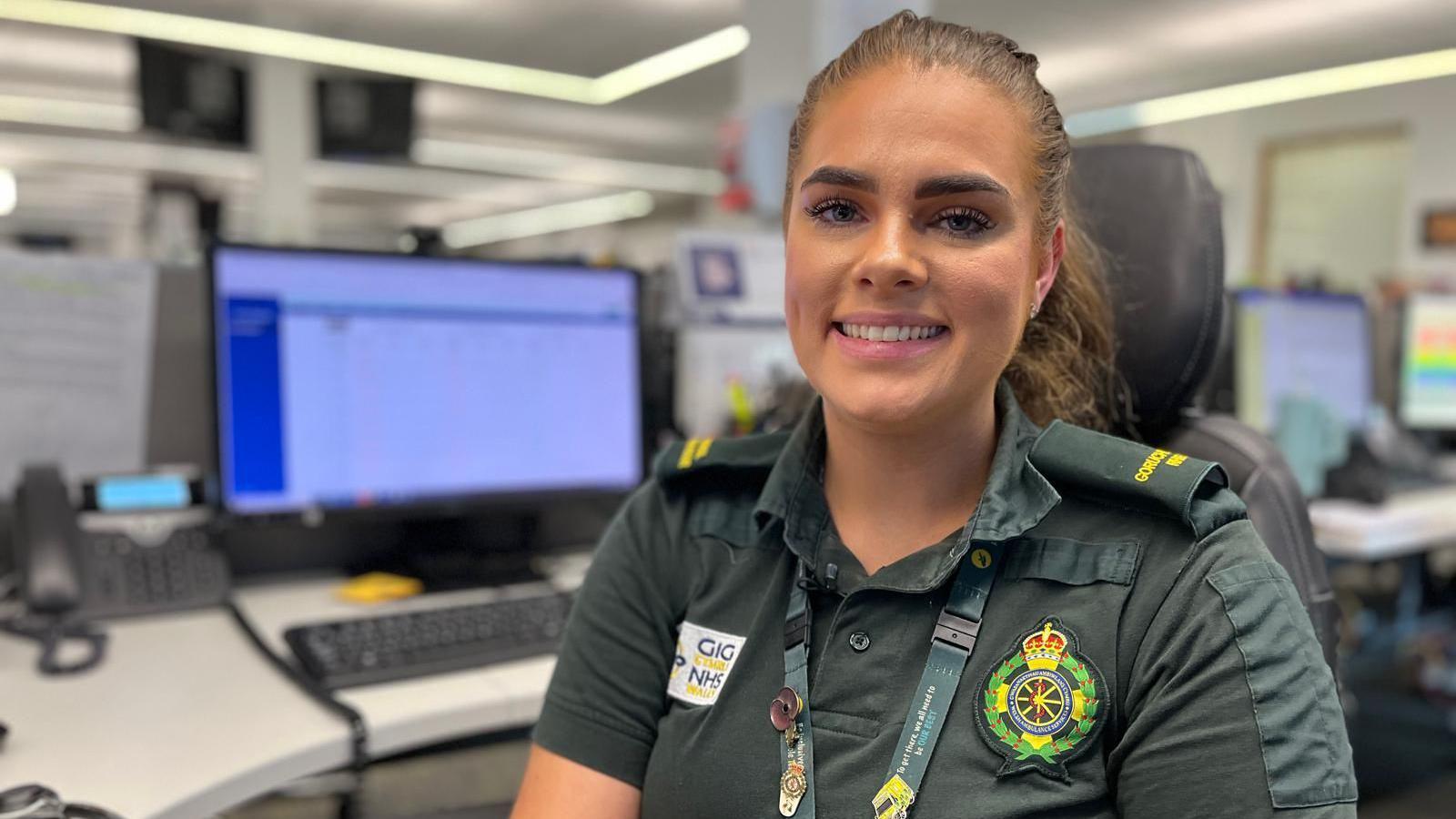 Abbie is wearing a green ambulance service uniform, with embroidered logos and a work lanyard. She has long dark hair tied back off her face and is smiling at the camera. She is sat in the control room at Cwmbran, with computer screens blurred in the background.
