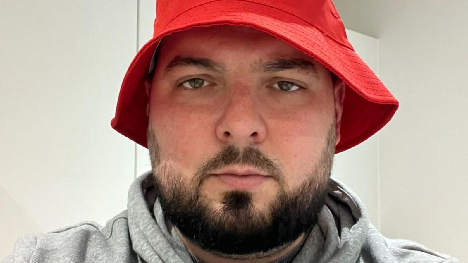 A man in a red bucket hat and grey hoodie looks at the camera.