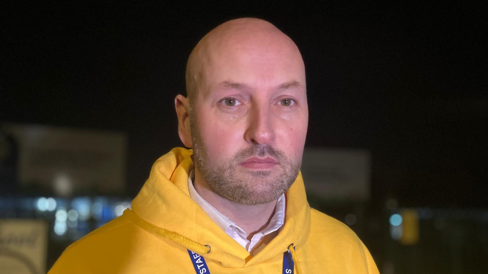 Headteacher of Queensway Primary School in Yeadon, Leeds, Mark Duce, stands outside of the school He wears a bright yellow hoodie. The school building is blurred behind him. It is nighttime.