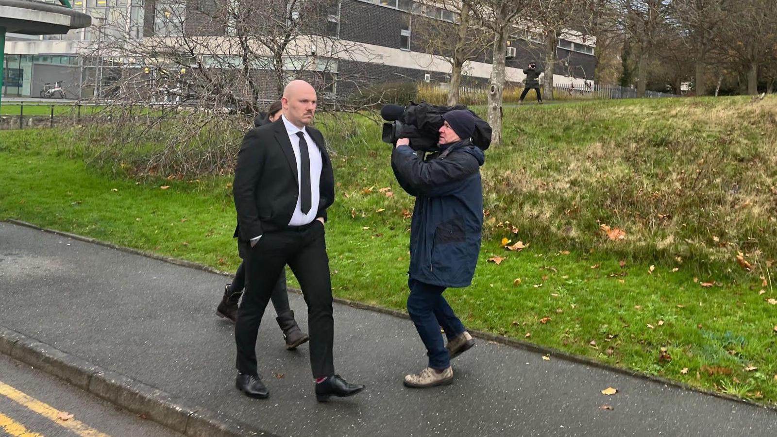 Miles Cross, mewn crys gwyn a siwt a thei du, yn gadael y llys, gan anwybyddu criw newyddion oedd yn ceisio ei holi