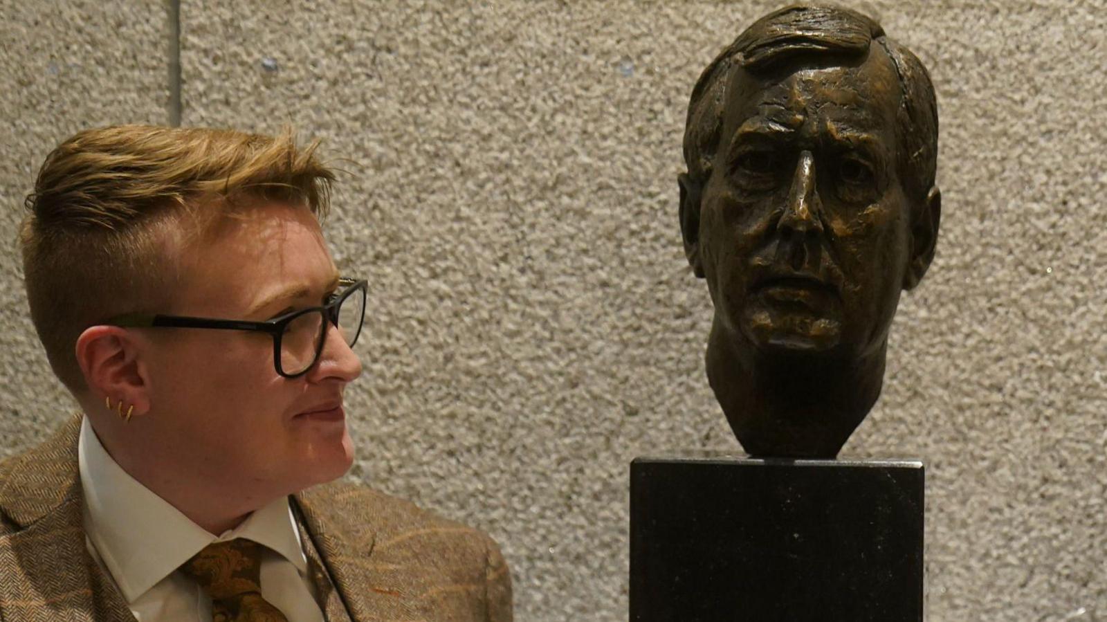 Vicky Trimble looking at a sculpture of the face of her late father Lord Trimble during its unveiling in Dublin in December 2023.  She is wearing black-rimmed glasses and a light-brown plaid three-piece suit and tie.