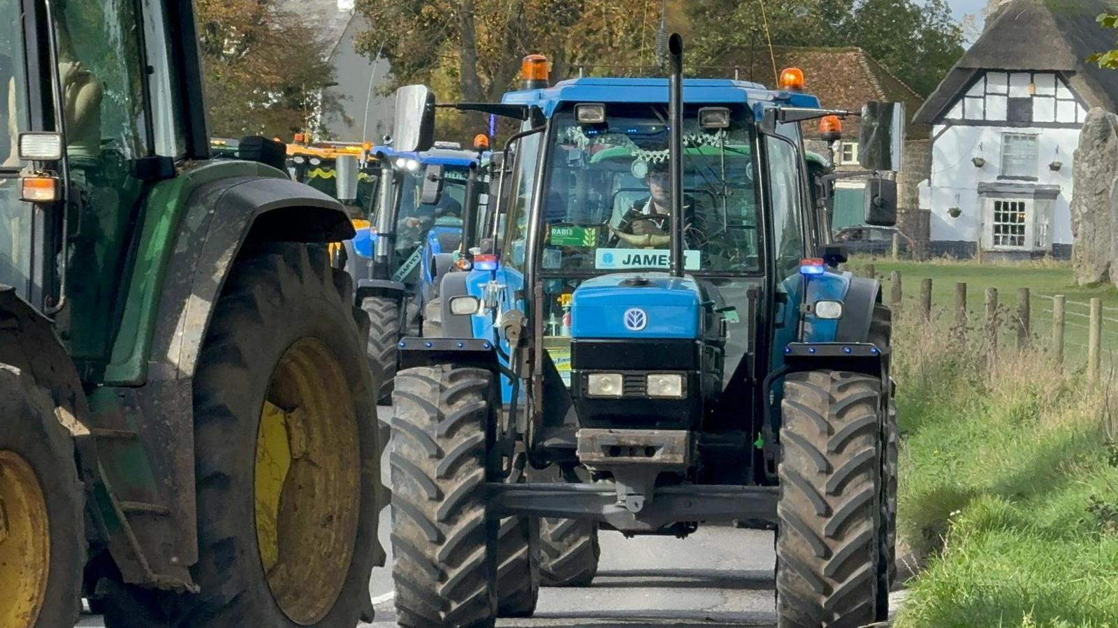 A close up of a blue tractor driven by a young man with a licence plate reading "James" in the cab window. It is part of a line of tractors passing through a country village. 