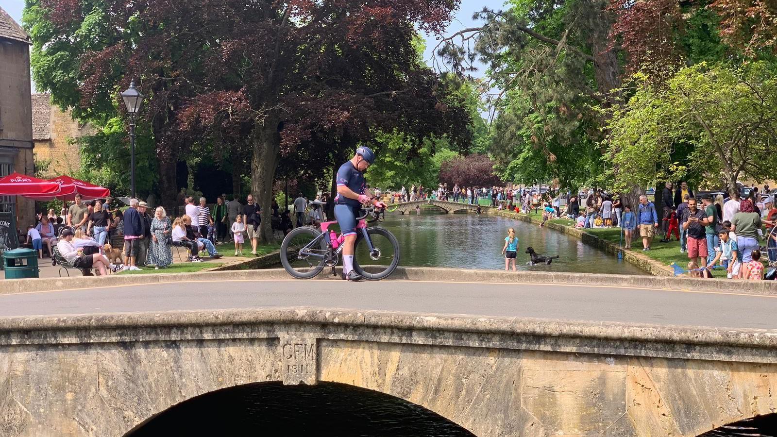 A small Cotswolds-stone bridge with a cyclist wearing Lycra stopped in the middle. The bridge goes over a picturesque river with crowds of people on either side sitting on the grass.