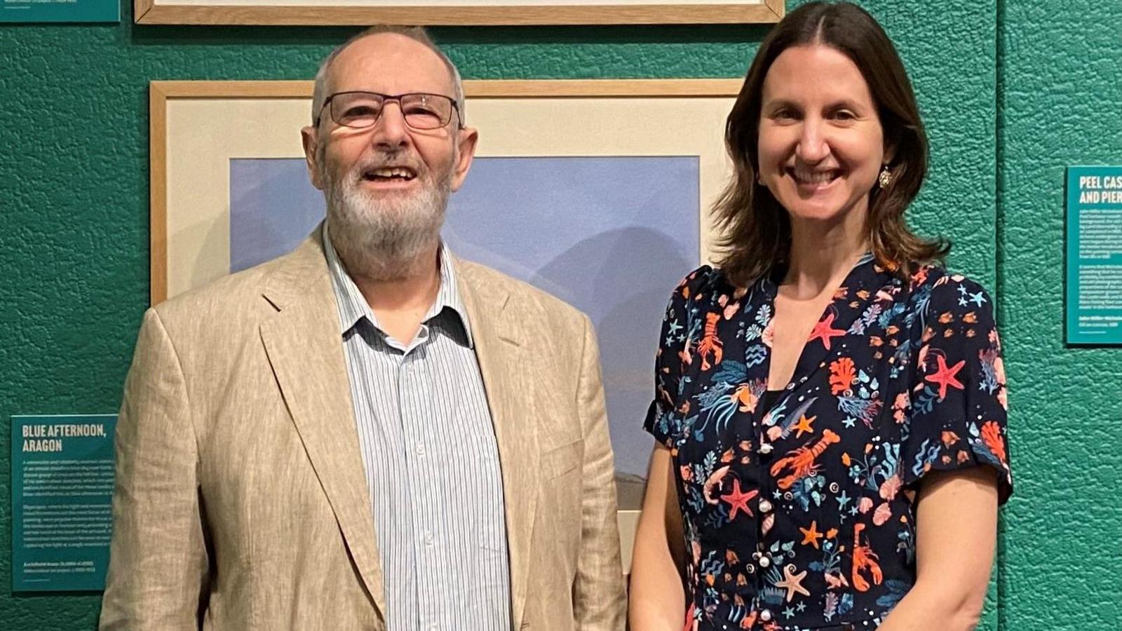 A man wearing a beige blazer and pale shirt standing alongside a woman wearing a black dress with colourful sea creature designs standing in against a green wall with paintings hanging on it. Both are smiling.