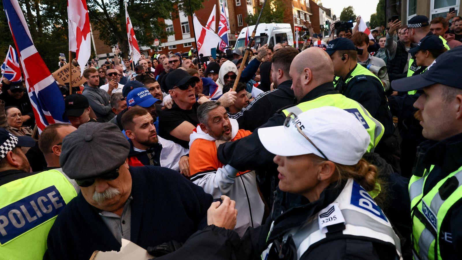 A group of people holding up Union flags and England flags standing against a line of police in uniform and fluorescent jackets. One man wearing and white jacket with a large orange stripe appears to be grappling with an officer