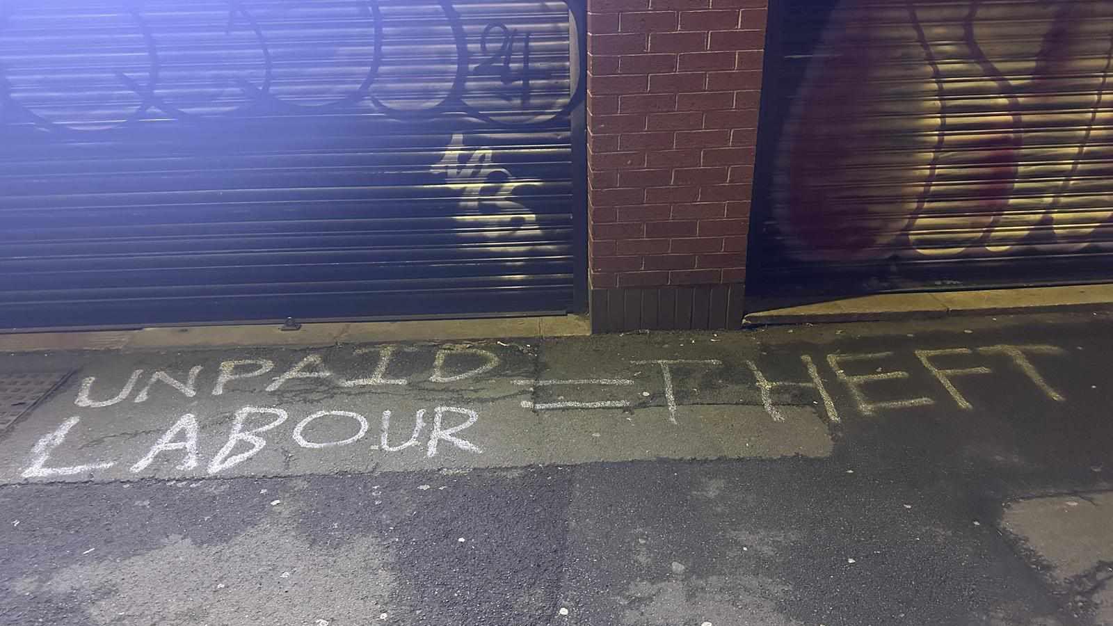 Graffiti sprayed in white paint on the ground outside one of the restaurants after it closed. The graffiti reads UNPAID LABOUR = THEFT