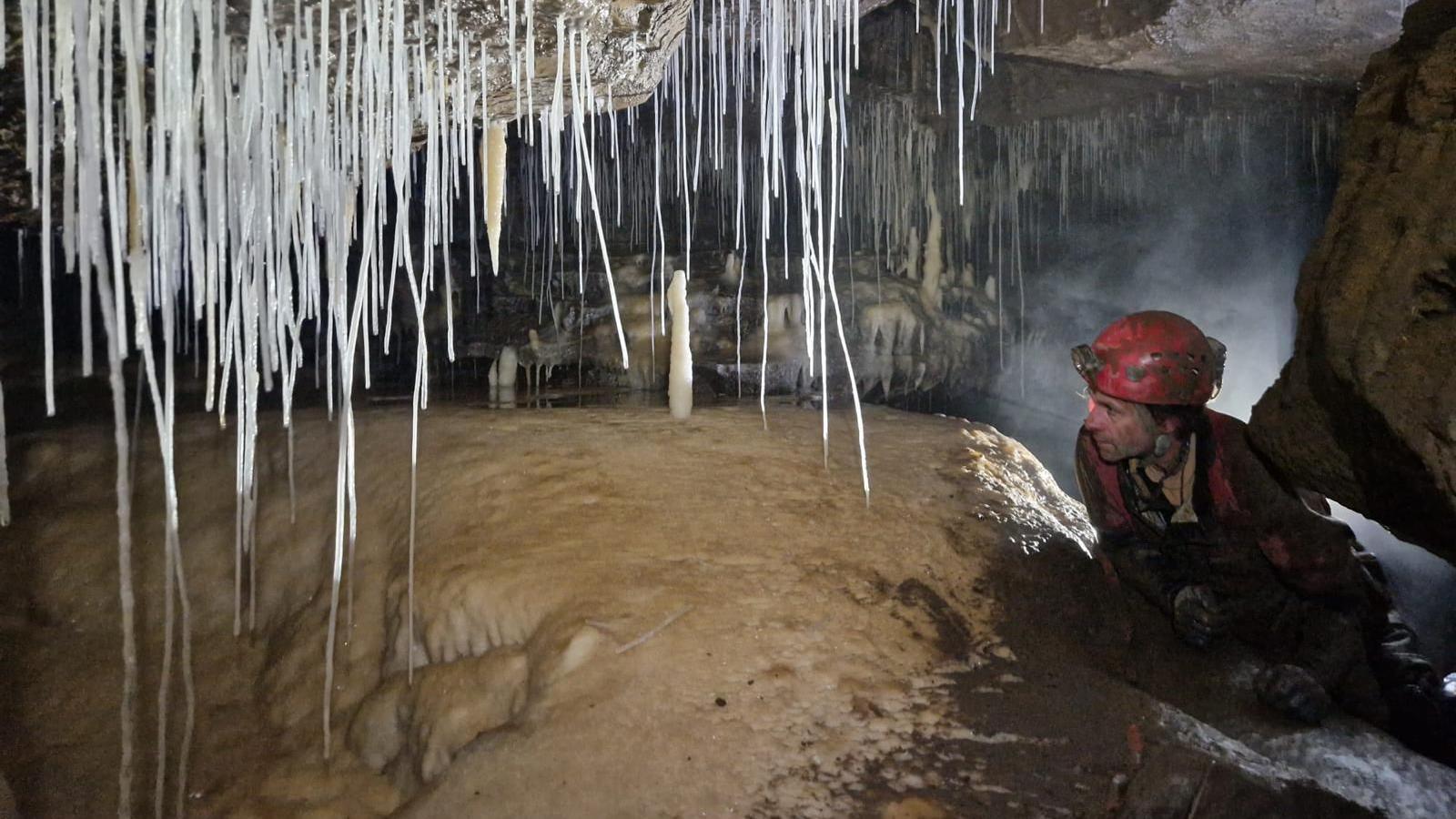 The photo shows a person in full caving gear, including a helmet, crawling through a narrow cave passage surrounded by long, thin stalactites hanging from the ceiling and stalagmites rising from the ground.