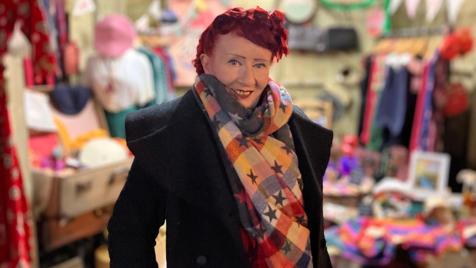 Elaine Duffy standing in front of her market stall. 
She has bright red hair and is wearing a long black coat with a multi-coloured scarf. 