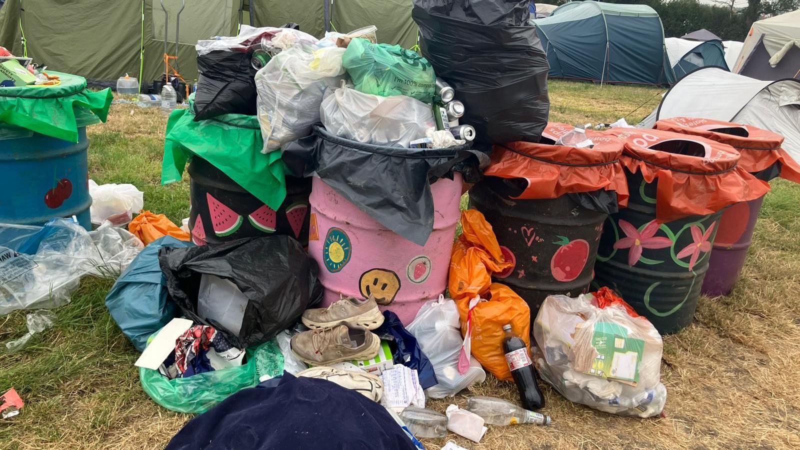 Several bins at a campsite at Glastonbury Festival. They are overflowing and there is rubbish on the floor around them. Lots of tents can be seen in the distance.