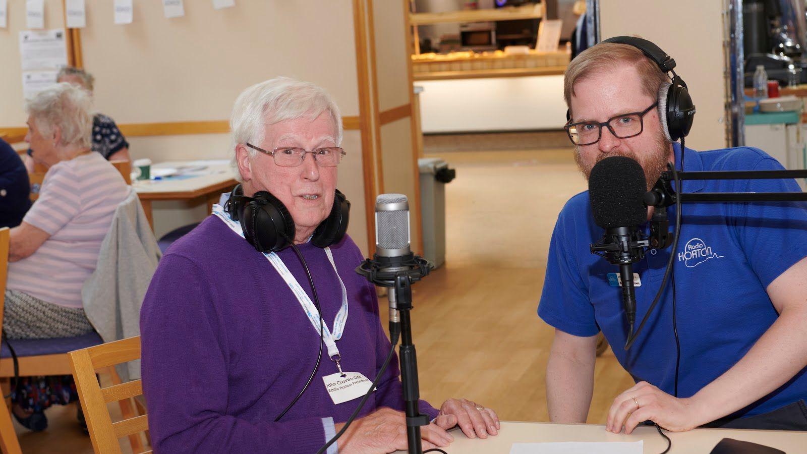 John Craven who has short, grey hair and is wearing glasses and a purple jumper sits next to a younger man in a blue t shirt - both have headphones and mics