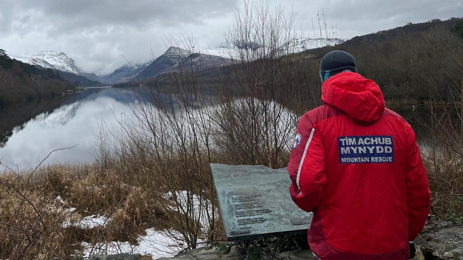 Un o wirfoddolwyr tim achub mynydd yn darllen arwydd o flaen y llun a'r mynyddoedd yn y cefndir. Mae eira ar y mynyddoedd