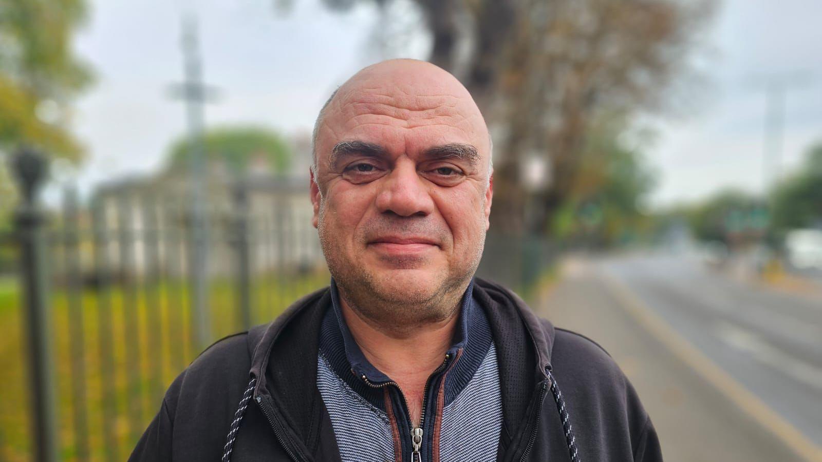 Mehmet Dalgic is looking directly at the camera, he has a black hoody and a t-shirt with a blue collar on. He has a bald head and stubble around his chin.