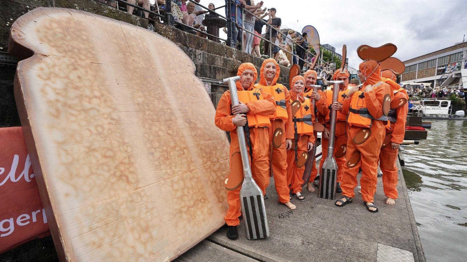 A group of people in bright orange waterproof outfits stand next to their home-made craft, which looks like a slice of bread. Two of them are holding paddles which are designed to look like forks. It is part of the Bristol Harbour Festival cardboard boat race.