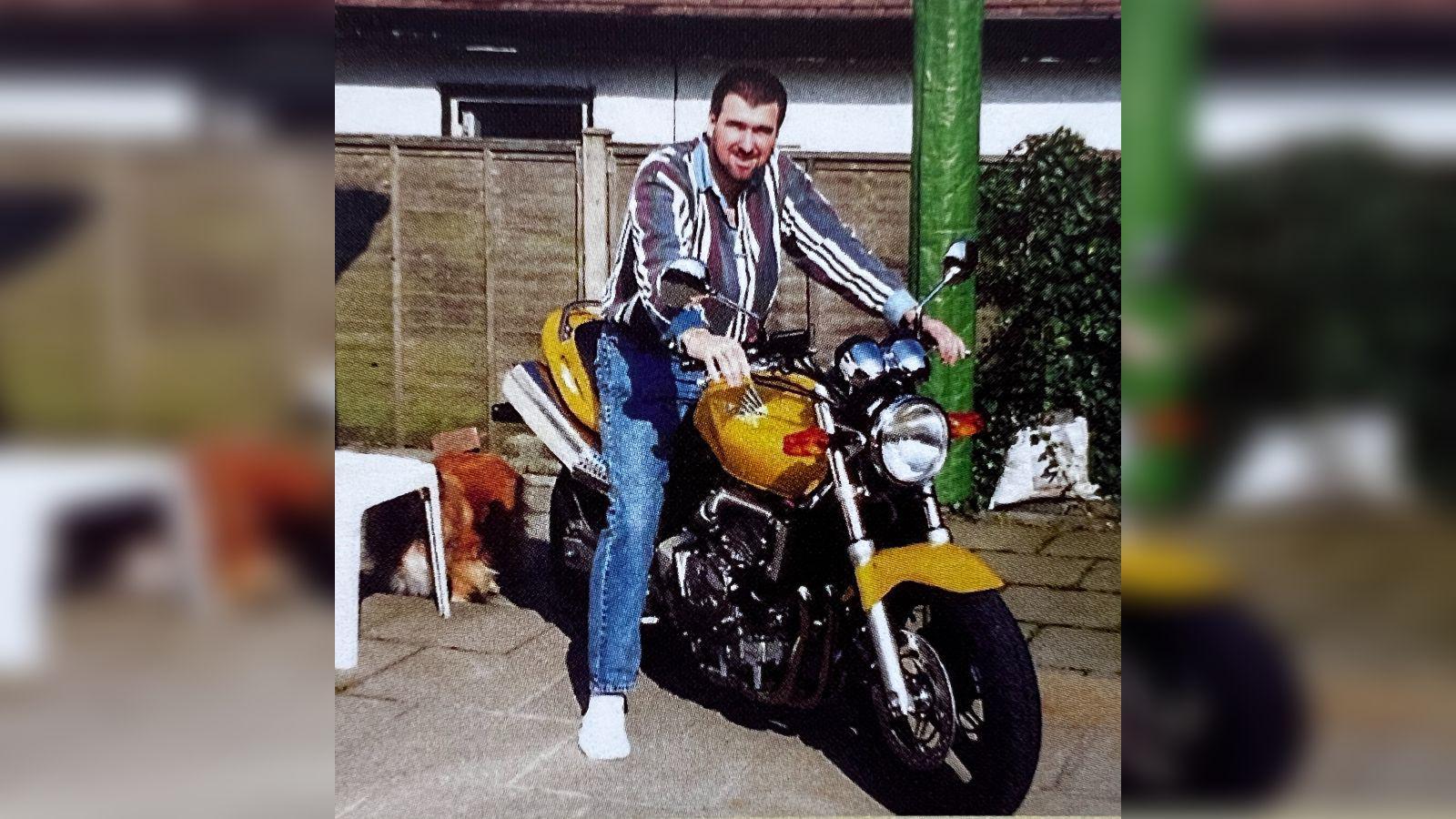 An old photograph of Mark Reeves standing astride motorbike with gold-coloured panels. He is wearing a striped shirt, blue jeans and white socks. The image looks typical of the 1990s. 