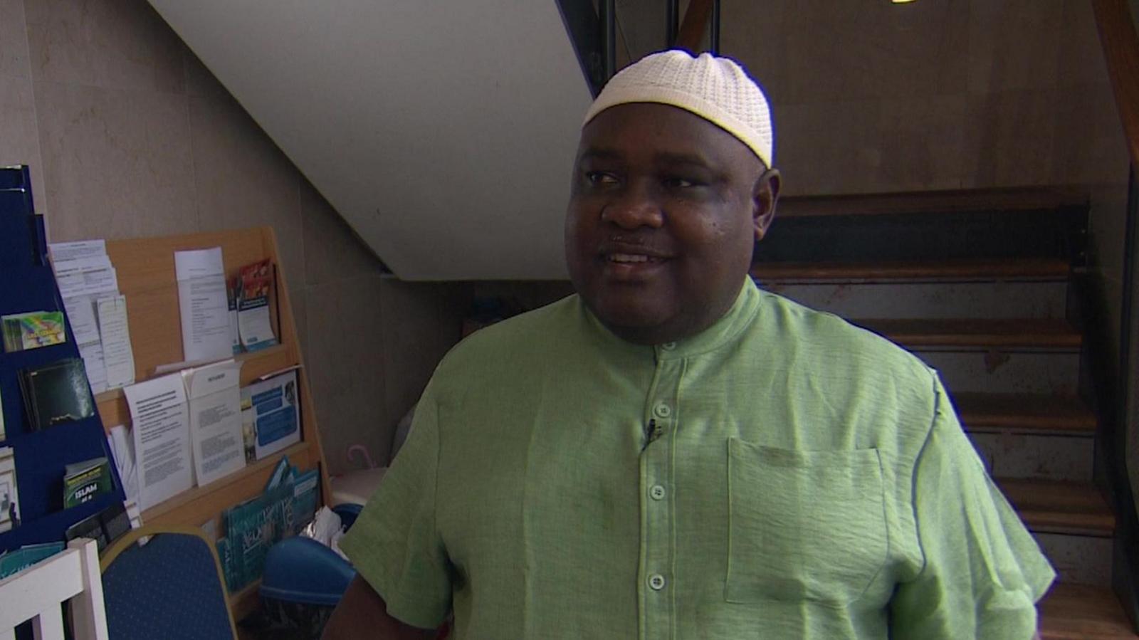 The centre director a mosque is stood in front of a flight of stairs smiling at the camera. He is wearing a green shirt.