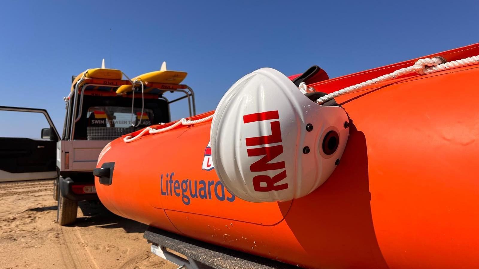 An orange inflatable rescue boat, with Lifeguards on the side, with a white helmet saying RNLI hanging off the side of it. The boat is a attached to a white 4x4 vehicle which has two yellow rescue boards on the roof