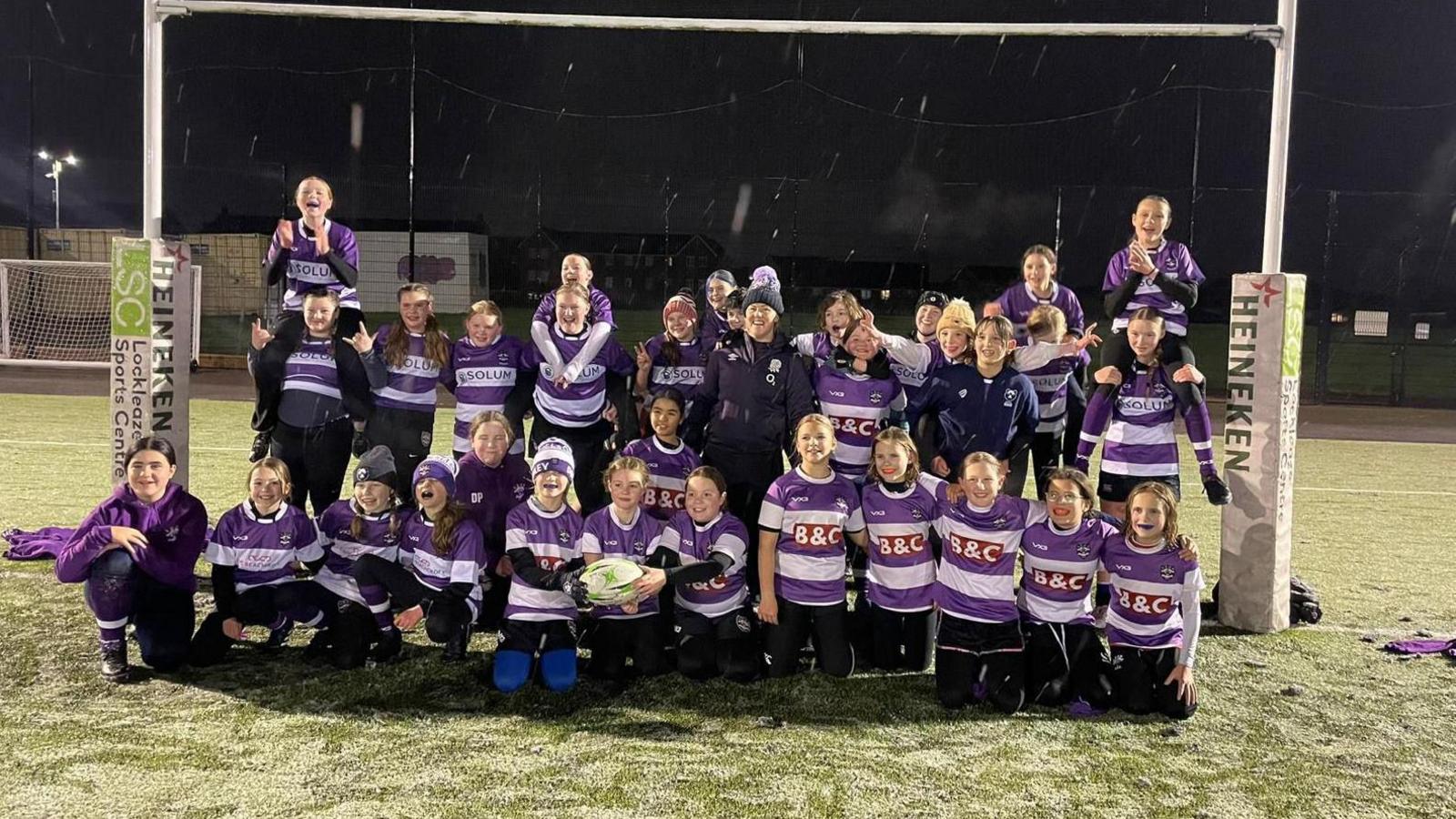 Marlie stands in the centre of a large group of girls, all of whom are wearing the purple and white striped rugby shirts for the team. They are standing between the goal posts on a floodlit pitch, and it is lightly snowing. 