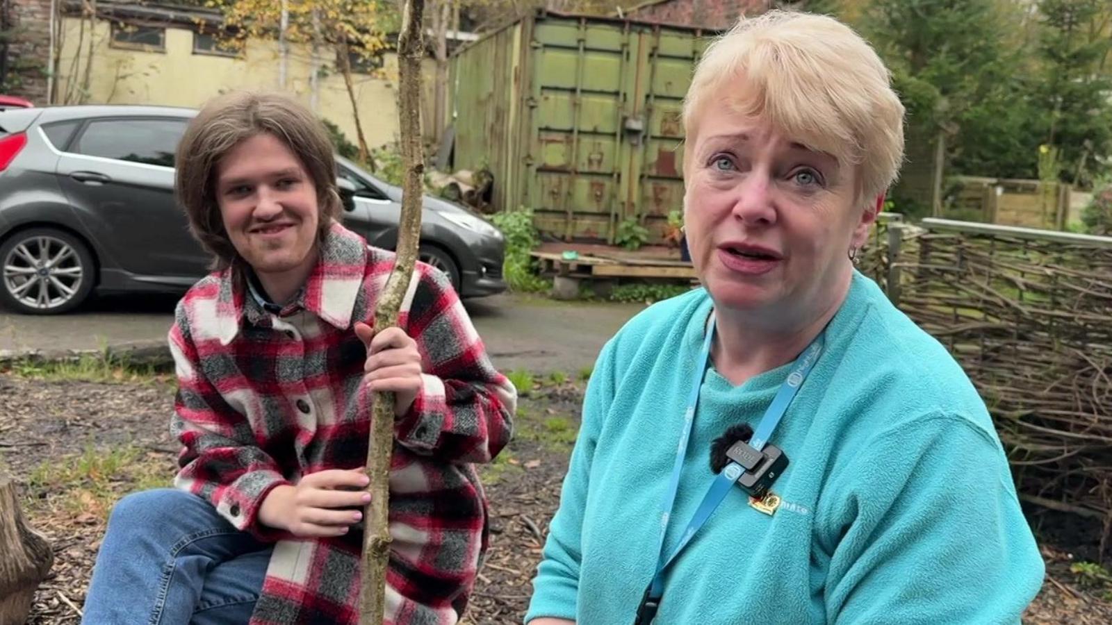 Adam is sitting in a checked shirt holding a stick, with Jane Wadsworth who has blonde hair and is wearing a light blue jumper. They are sat on logs next to a small camp fire.