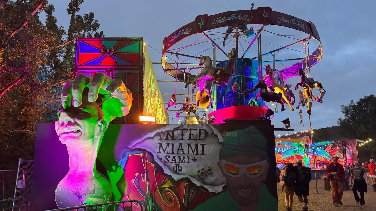 Unfairground at Glastonbury Festival. There is a carousel of skeletal horses high in the air while on the left is a grimacing head gripped from above by a large hand.
