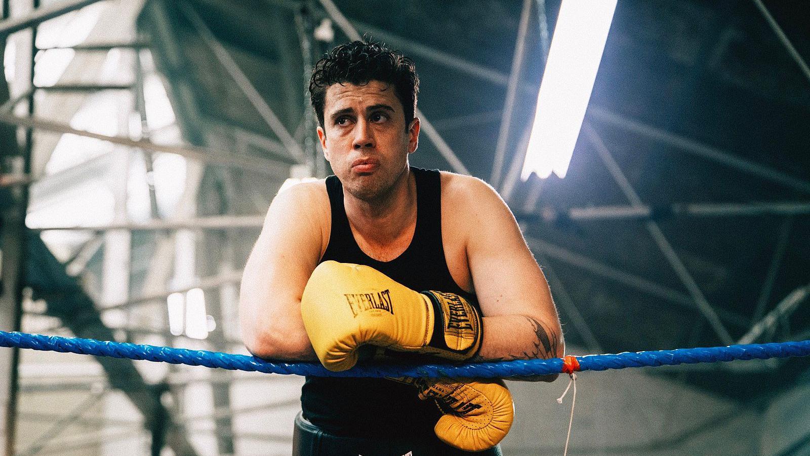 Toby Kebell wearing bright yellow everlast boxing gloves. He leans on blue rope of a boxing ring and wears a black vest. He has brown hair and stubble. He looks over to the corner. 