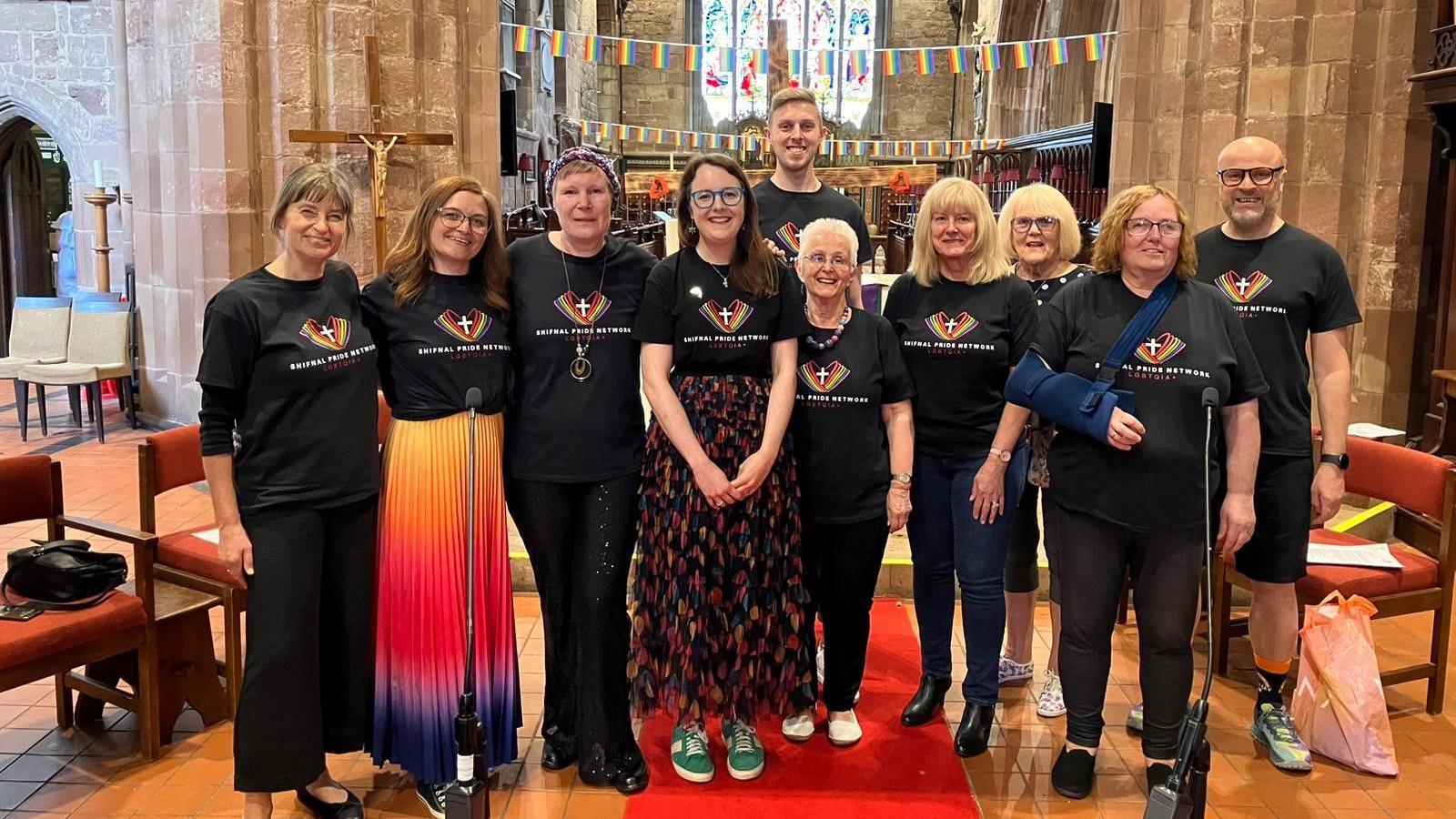 A group of people are inside a church. They are dressed in black T-shirts with a rainbow and a cross on them. Behind them, rainbow flags are strung up on banners.