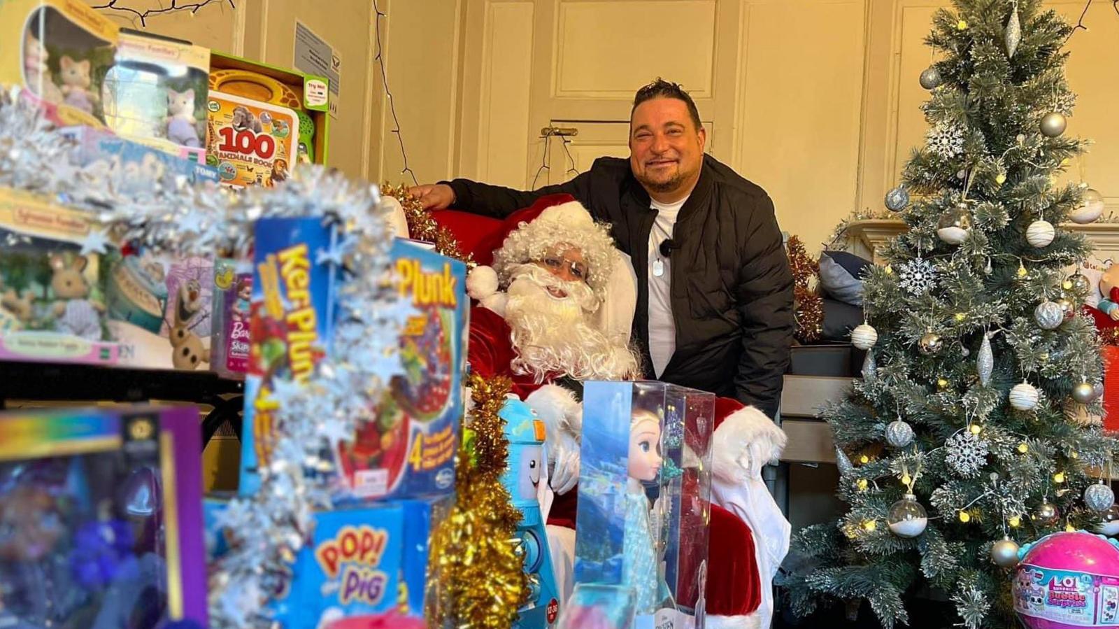 Wayne Bavin smiles at the camera while standing amongst a collection of toys. A Christmas tree also sits next to him. He has short dark hair and is wearing a black coat with a white t-shirt underneath.