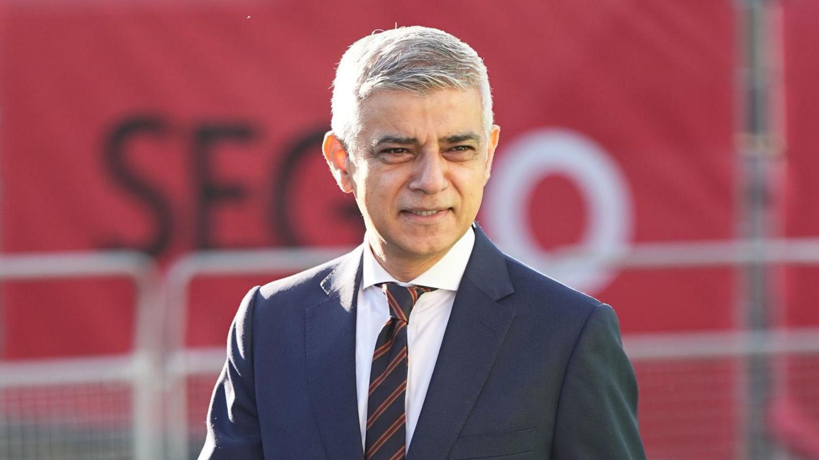 Sadiq Khan, a man with grey hair and brown eyes, wearing a dark suit and tie and white shirt, looks as crowds out of shot as he walks along in London